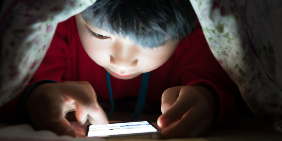 Un jeune garçon caché sous sa couverture avec un téléphone, une image illustrant l’usage smartphone des enfants.