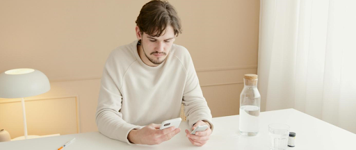 Un homme assis à une table, smartphone à la main et des médicaments dans l’autre. Un homme assis à une table, smartphone à la main et des médicaments dans l’autre.