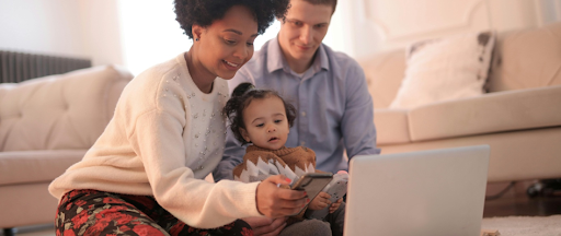 Une mère et un père assis à ras le sol avec leur enfant, postés devant un PC et un smartphone. Une mère et un père assis à ras le sol avec leur enfant, postés devant un PC et un smartphone.