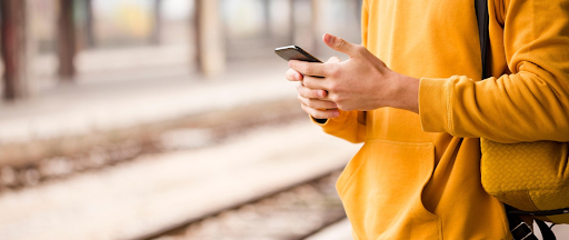 Une personne vêtue d’un pull jaune, debout dans un quai de gare et smartphone à la main. Une personne vêtue d’un pull jaune, debout dans un quai de gare et smartphone à la main.