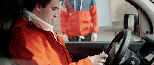 Un homme assis à son volant, tenant son smartphone dans les mains pendant qu’un homme l’attend à l’extérieur.
