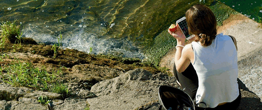 Une jeune femme assise au bord d’un point d’eau, en train de consulter son smartphone.